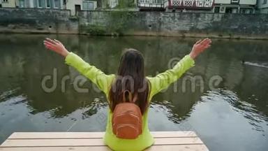 穿着<strong>黄</strong>色夹克的长发女人，背包坐在<strong>河边</strong>或池塘的木墩上，挥舞着双手。 传统